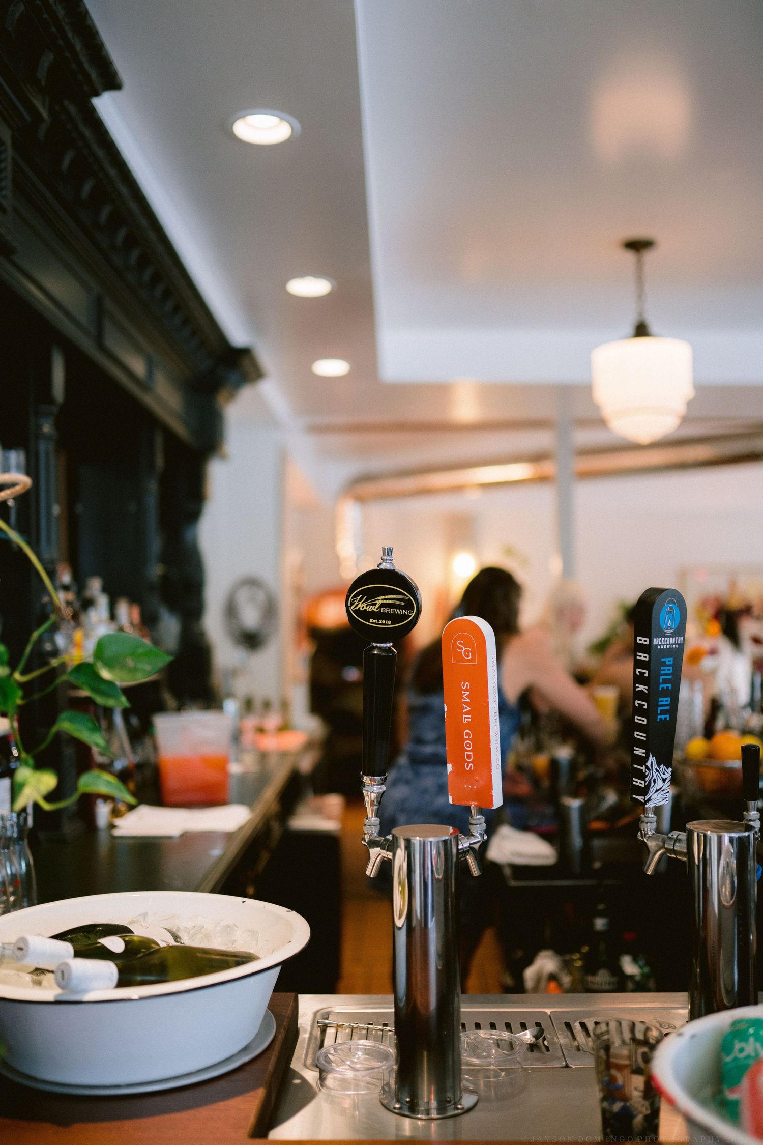 Row of beer taps at a bar counter with various draft handles and bottles in the background.