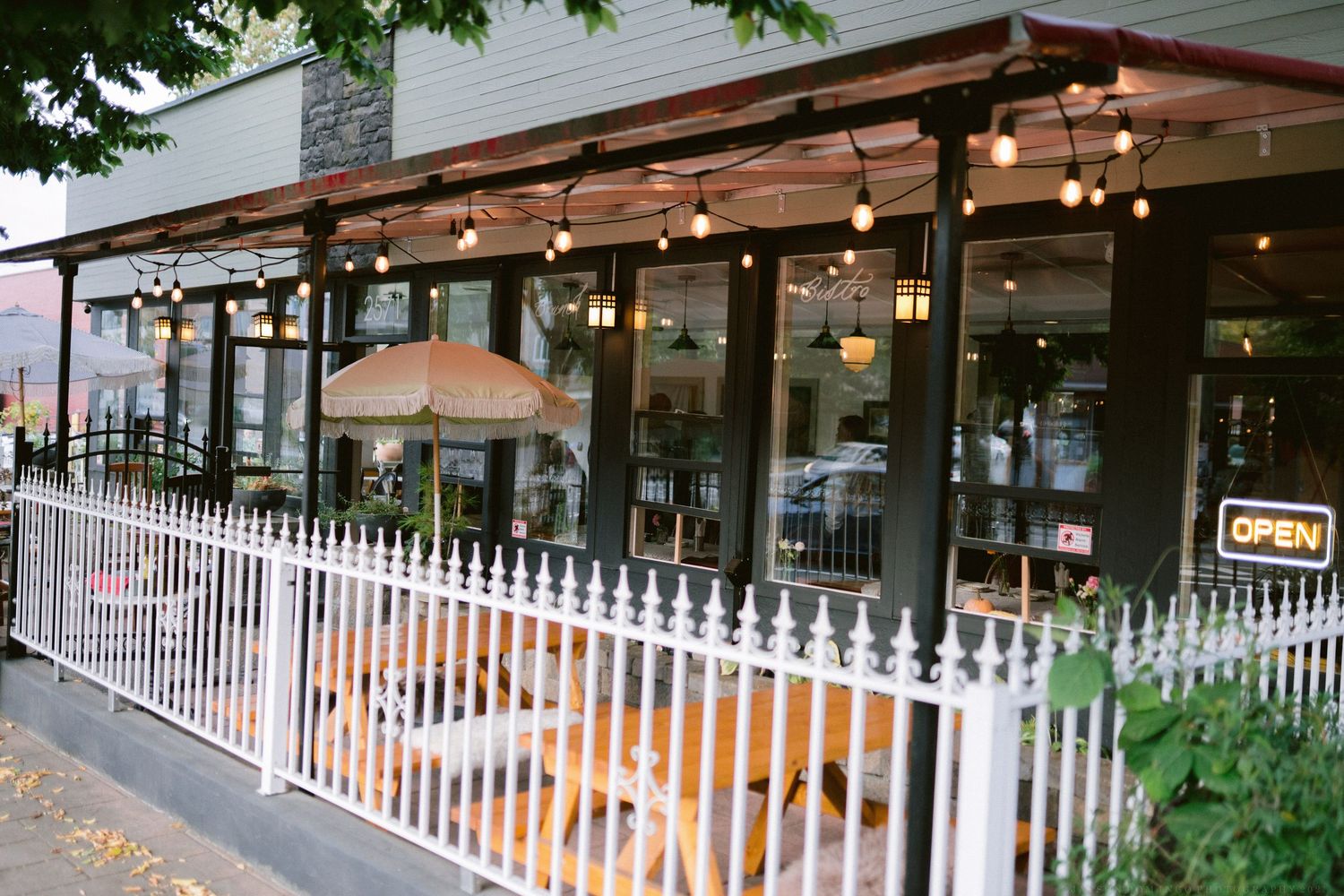 Restaurant patio with white picket fence and wooden pergola decorated with string lights and outdoor seating.