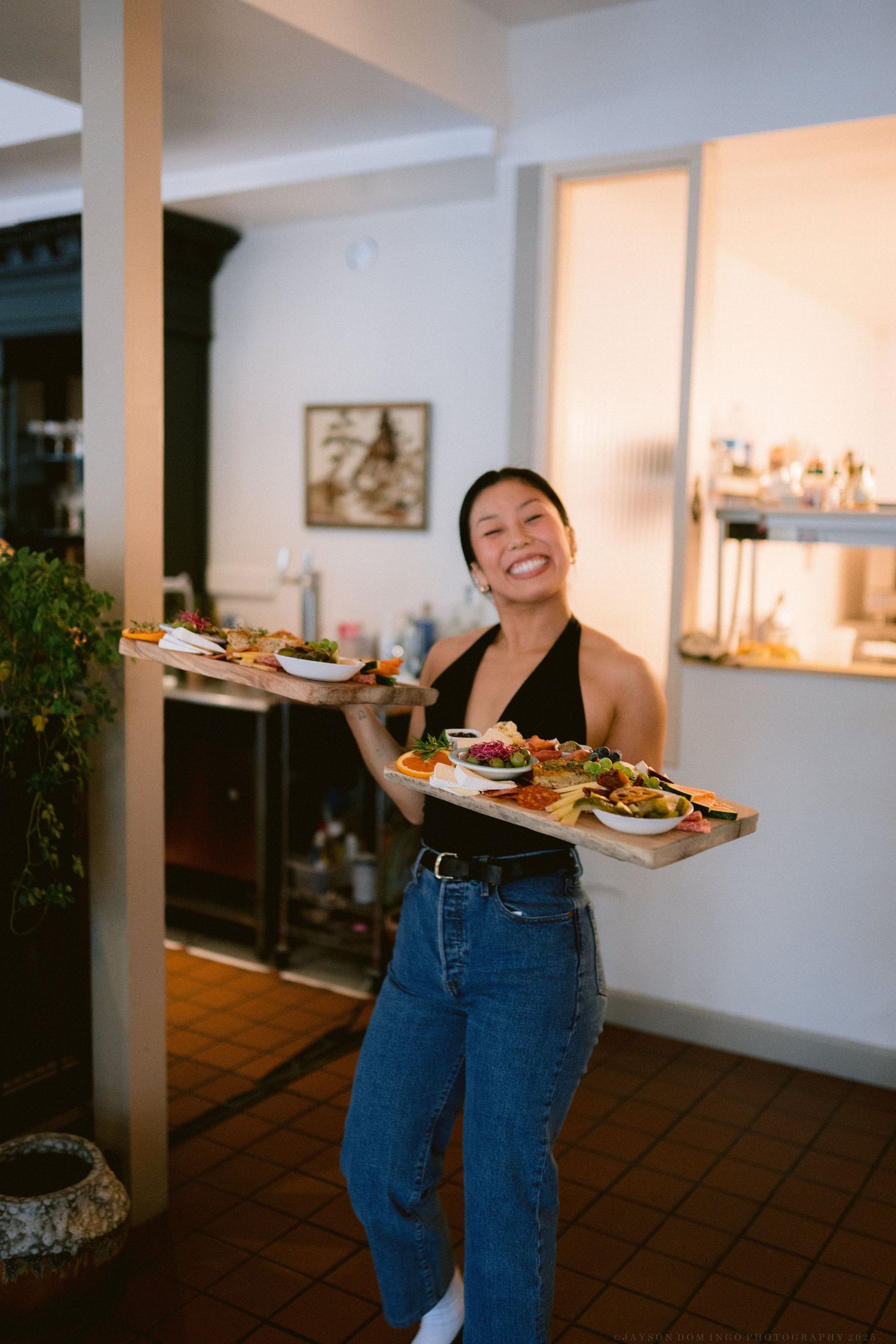 A person in jeans laughs joyfully while holding plates of food in a home dining area.