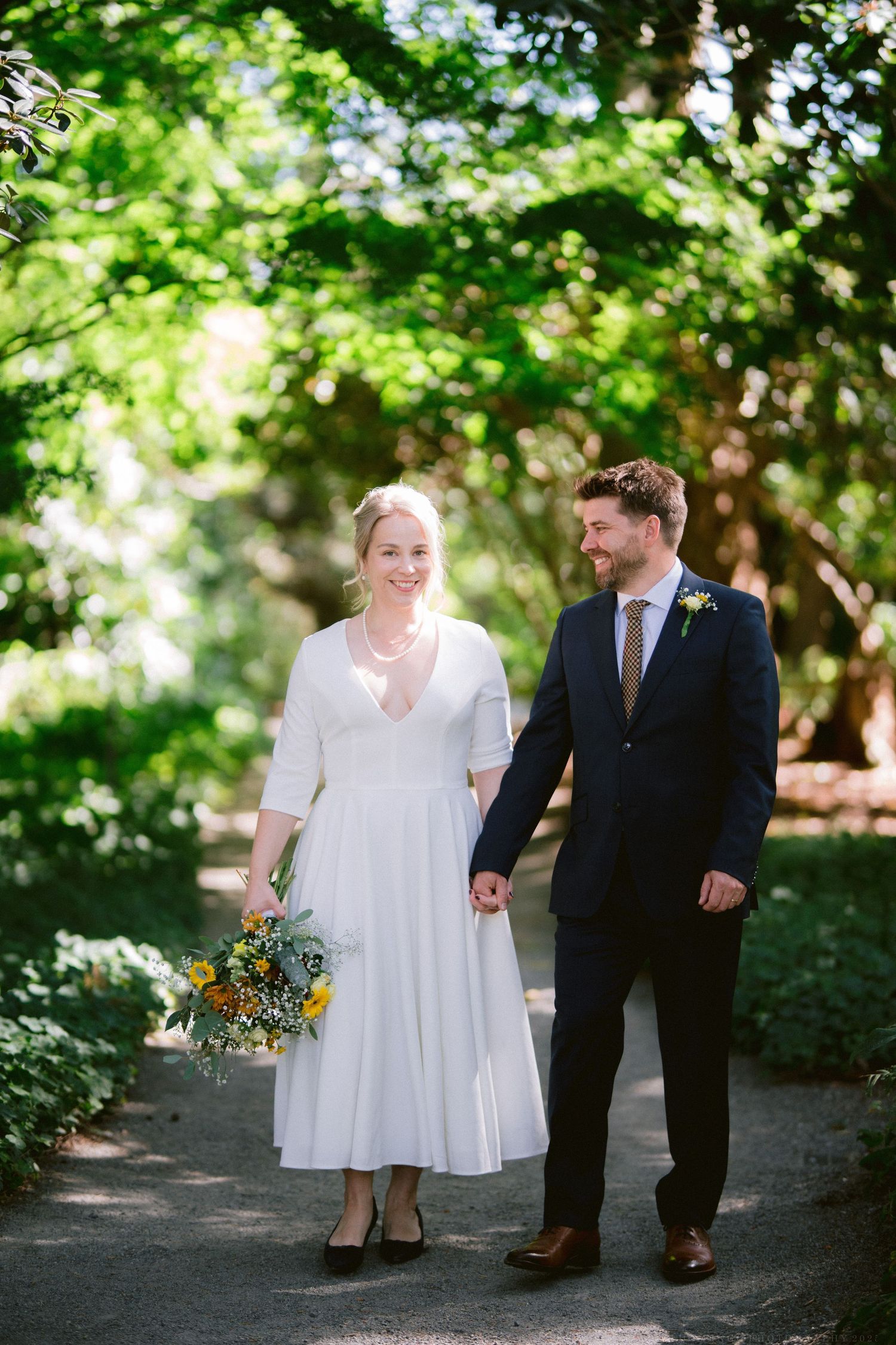 Newlyweds walk hand in hand along a shaded garden path on their wedding day.