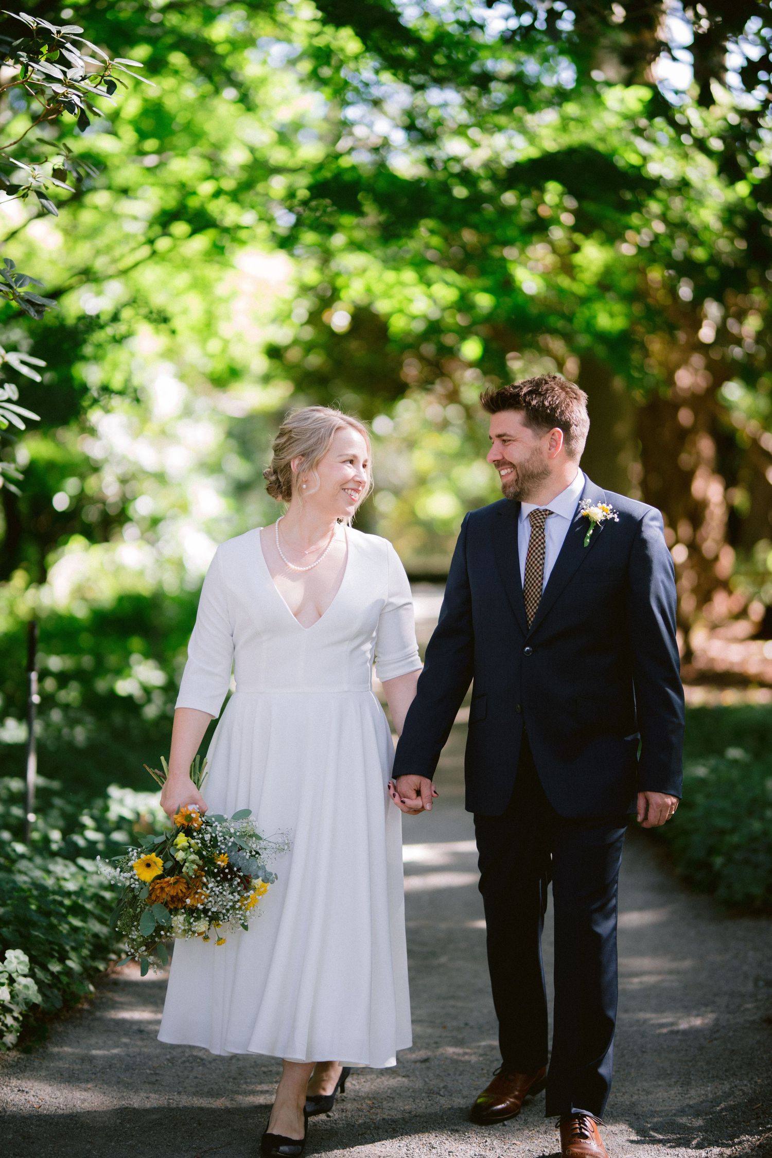 A romantic wedding couple walking together on a tree-lined path in a lush garden setting.