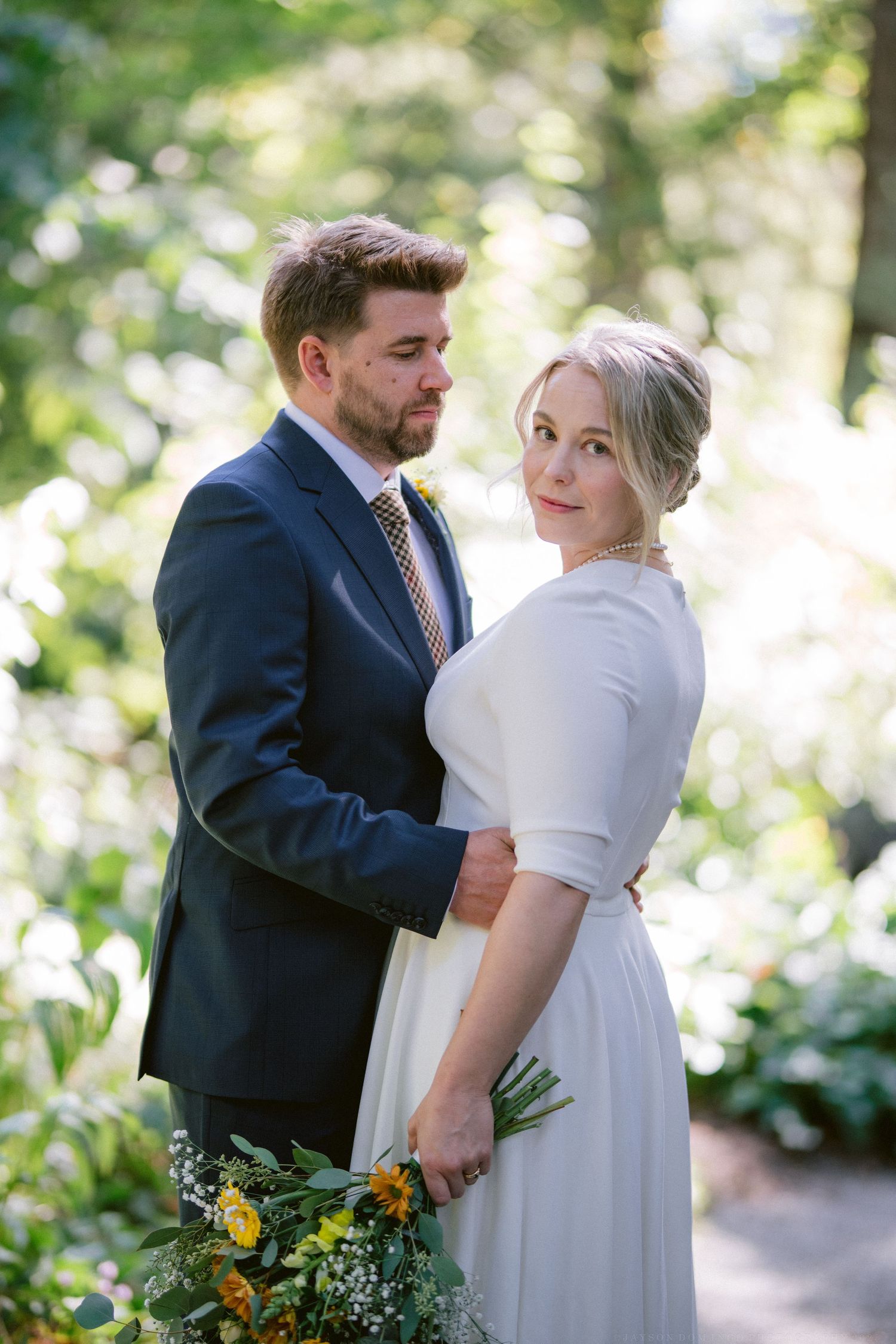 A tender moment between newlyweds in a sunlit garden, with the bride in white and groom in navy.