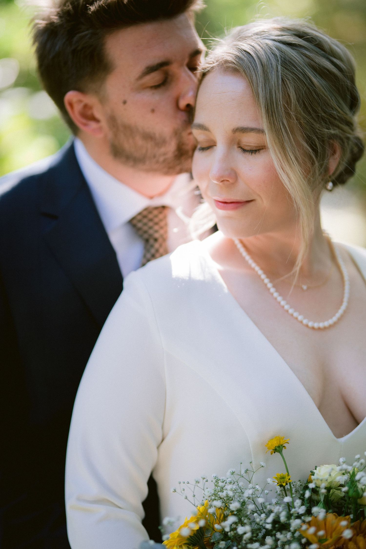 Intimate wedding portrait showing couple embracing outdoors with sunlit foliage in the background.