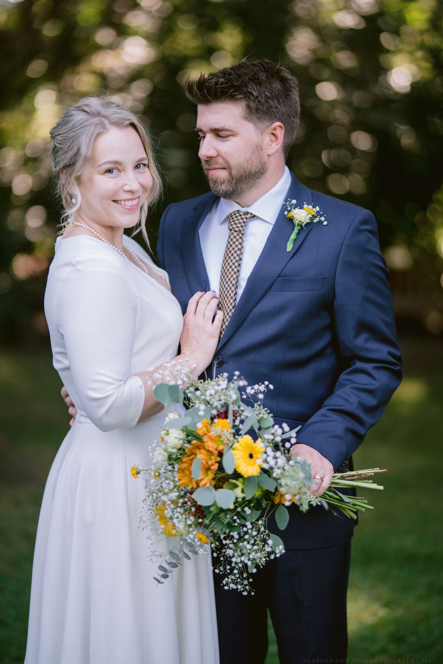 Couple in formal attire holding yellow and white floral bouquet outdoors.