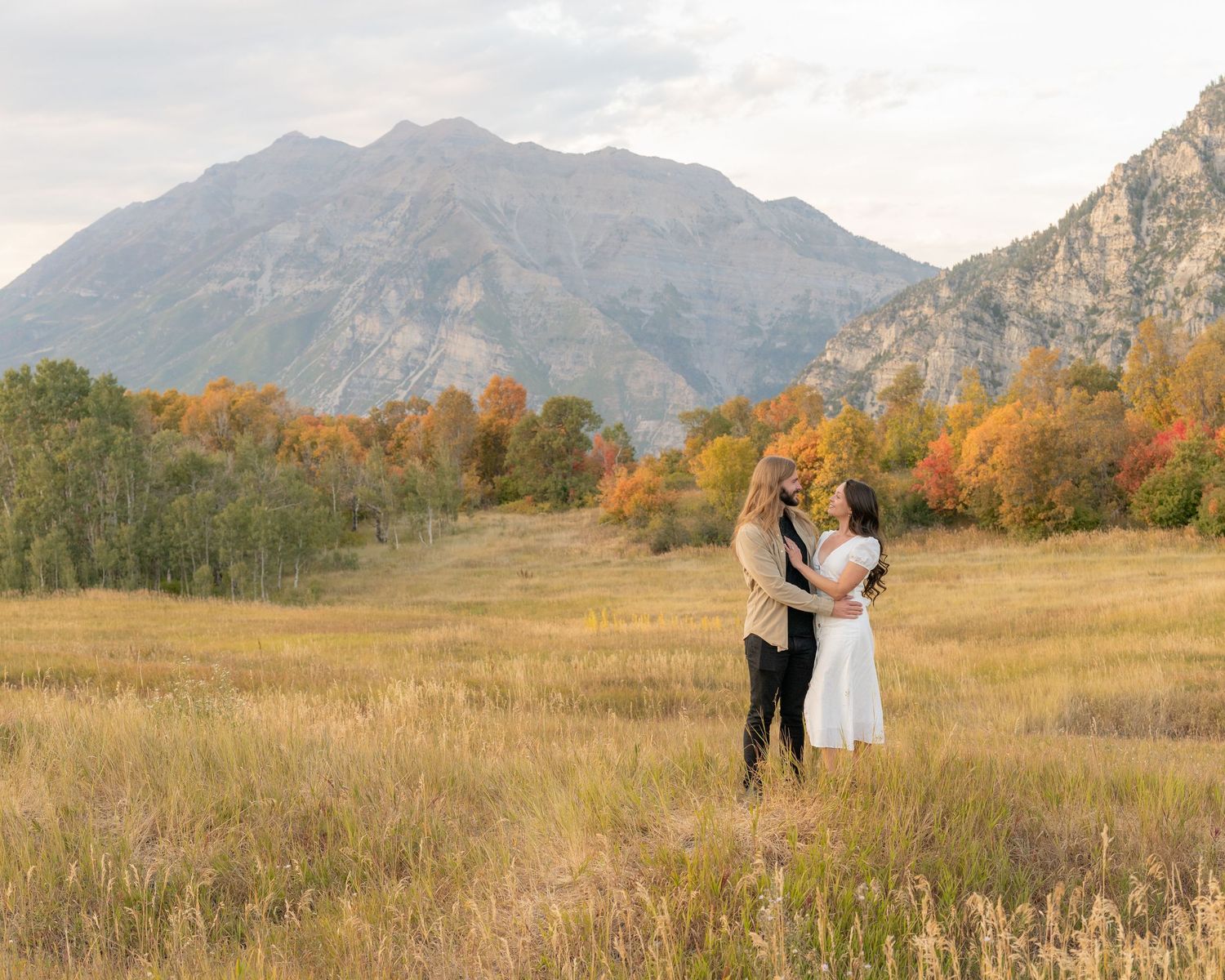 A couple poses together in a scenic mountain meadow with golden autumn grass and dramatic mountain peaks in the background.