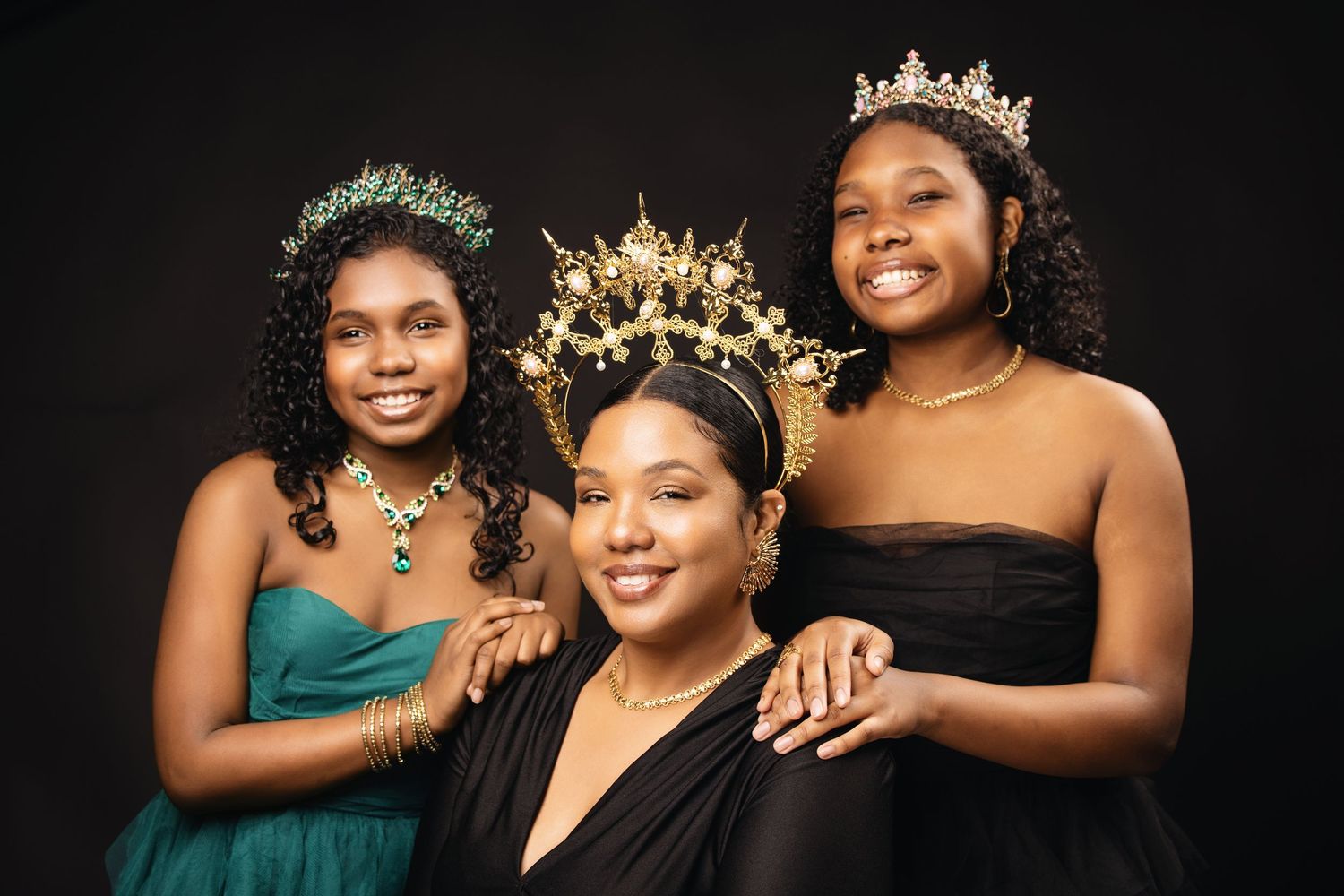 Three women wearing elegant evening gowns and crowns pose together for a formal portrait against a dark background.