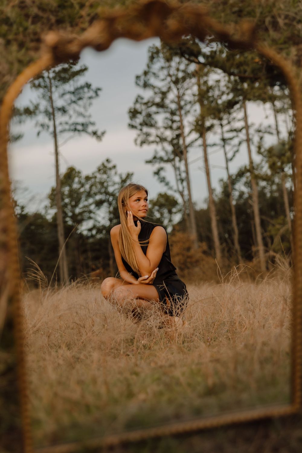 A woman in a black dress sits thoughtfully in tall golden grass with pine trees in the background during sunset.