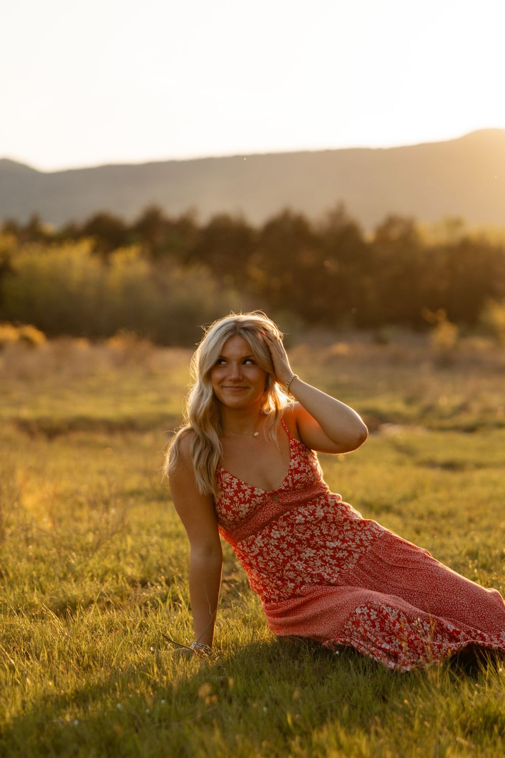 A woman in a red floral dress reclines in golden sunlit grass during sunset.
