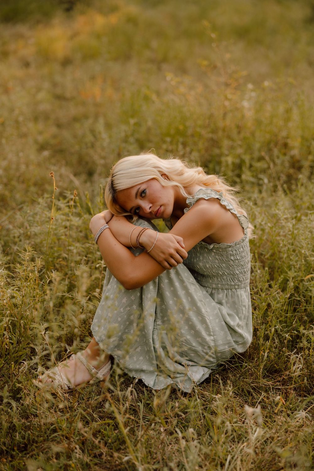 A person in a light dress sits curled up in a grassy meadow during golden hour.
