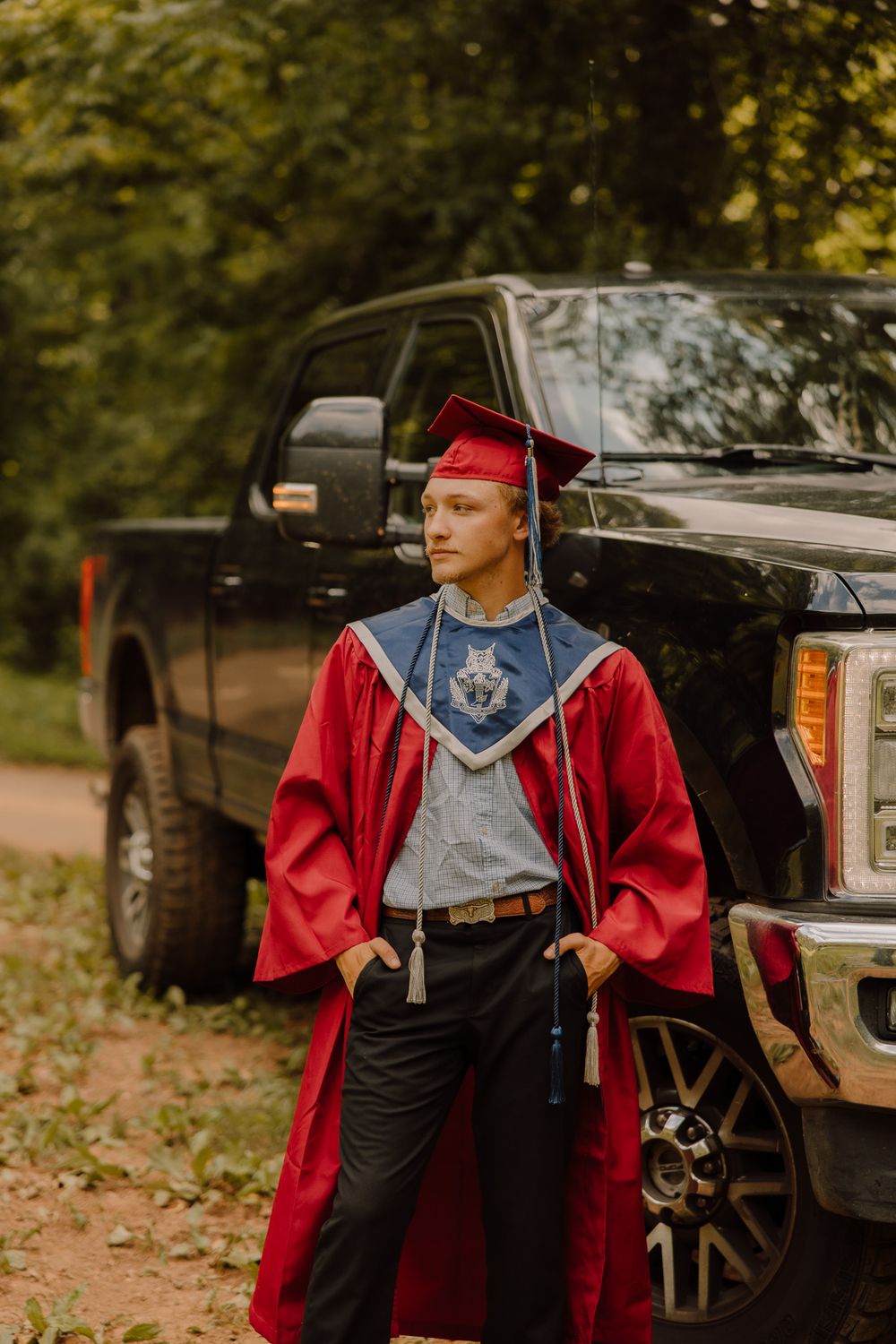 Person in a red graduation gown and black cap stands in front of a parked vehicle on a fall day.