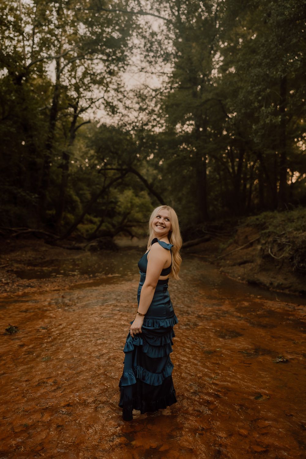 A woman in a flowing blue dress poses on a rustic dirt path surrounded by trees at sunset.