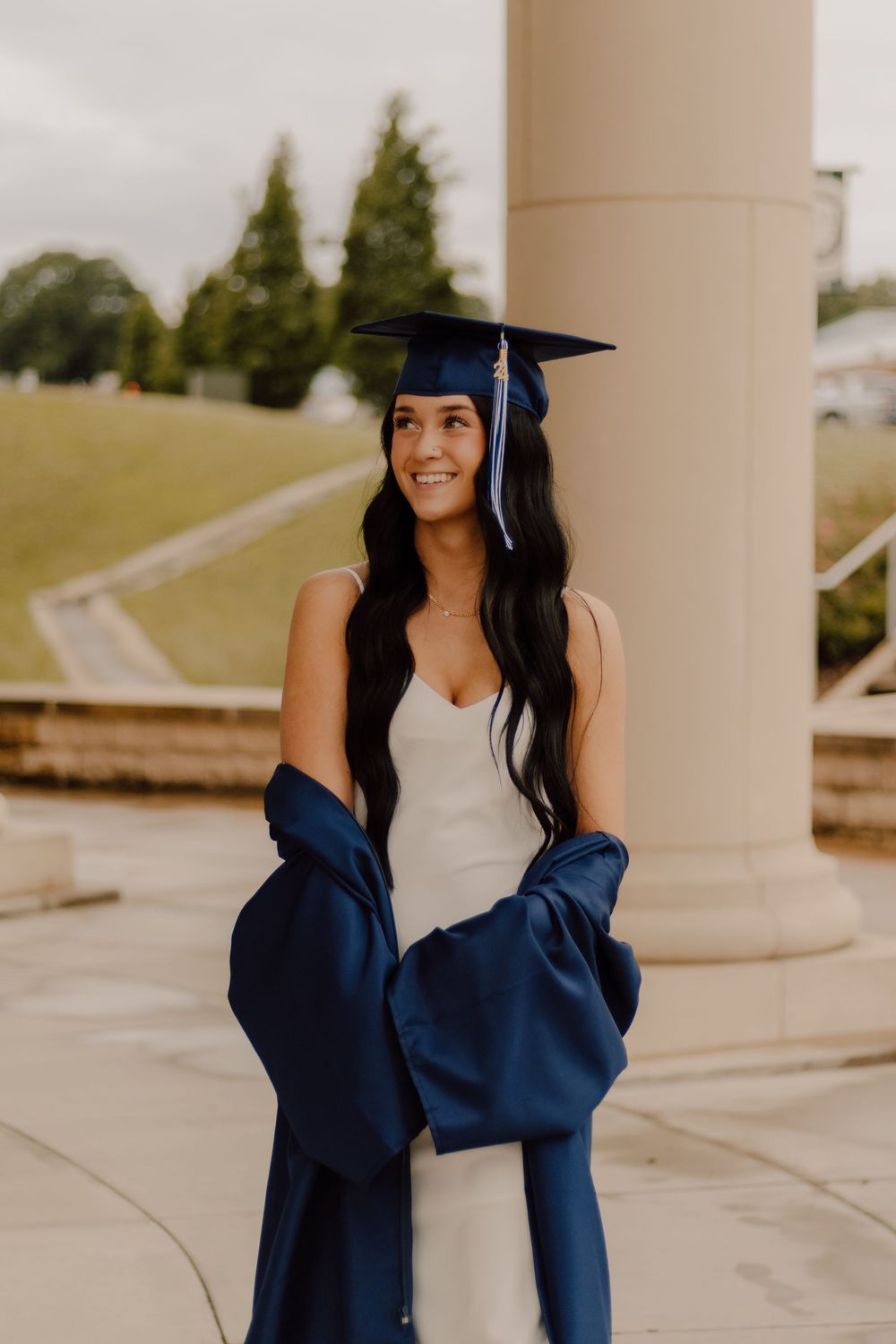 Graduate in blue cap and gown poses outdoors by white column in cream dress on sunny day.