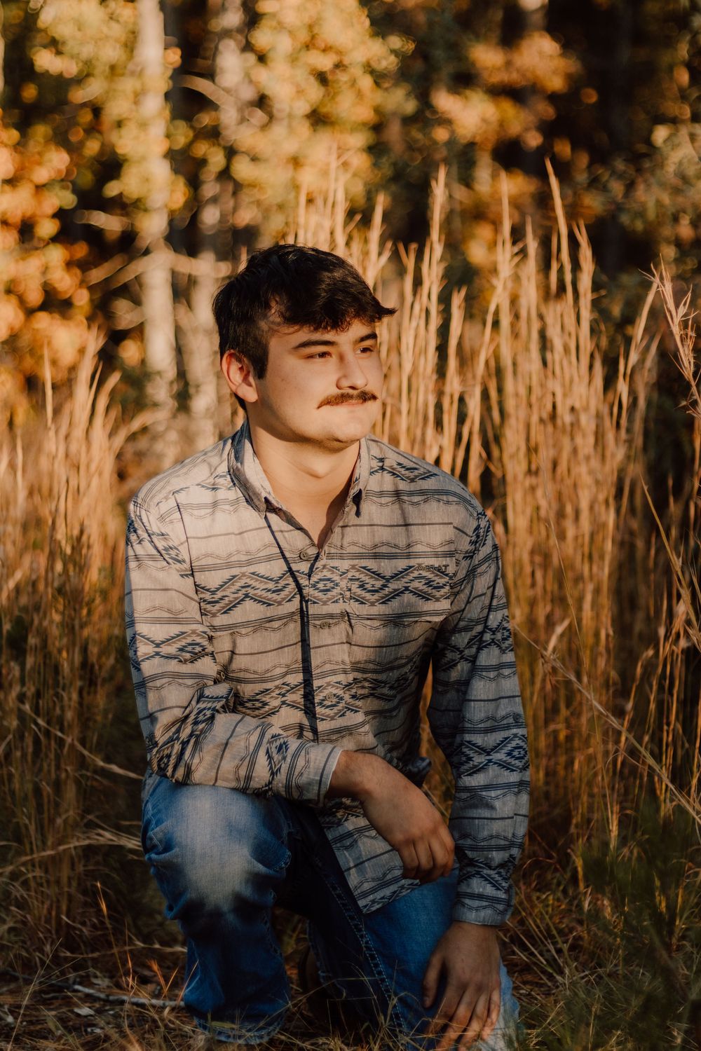 A young person in a patterned sweater sits in tall golden grass at sunset, creating a warm autumn portrait series.