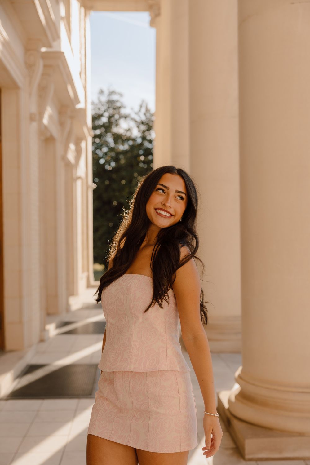 A woman in a pink two-piece outfit smiles while standing between elegant white columns in warm sunlight.
