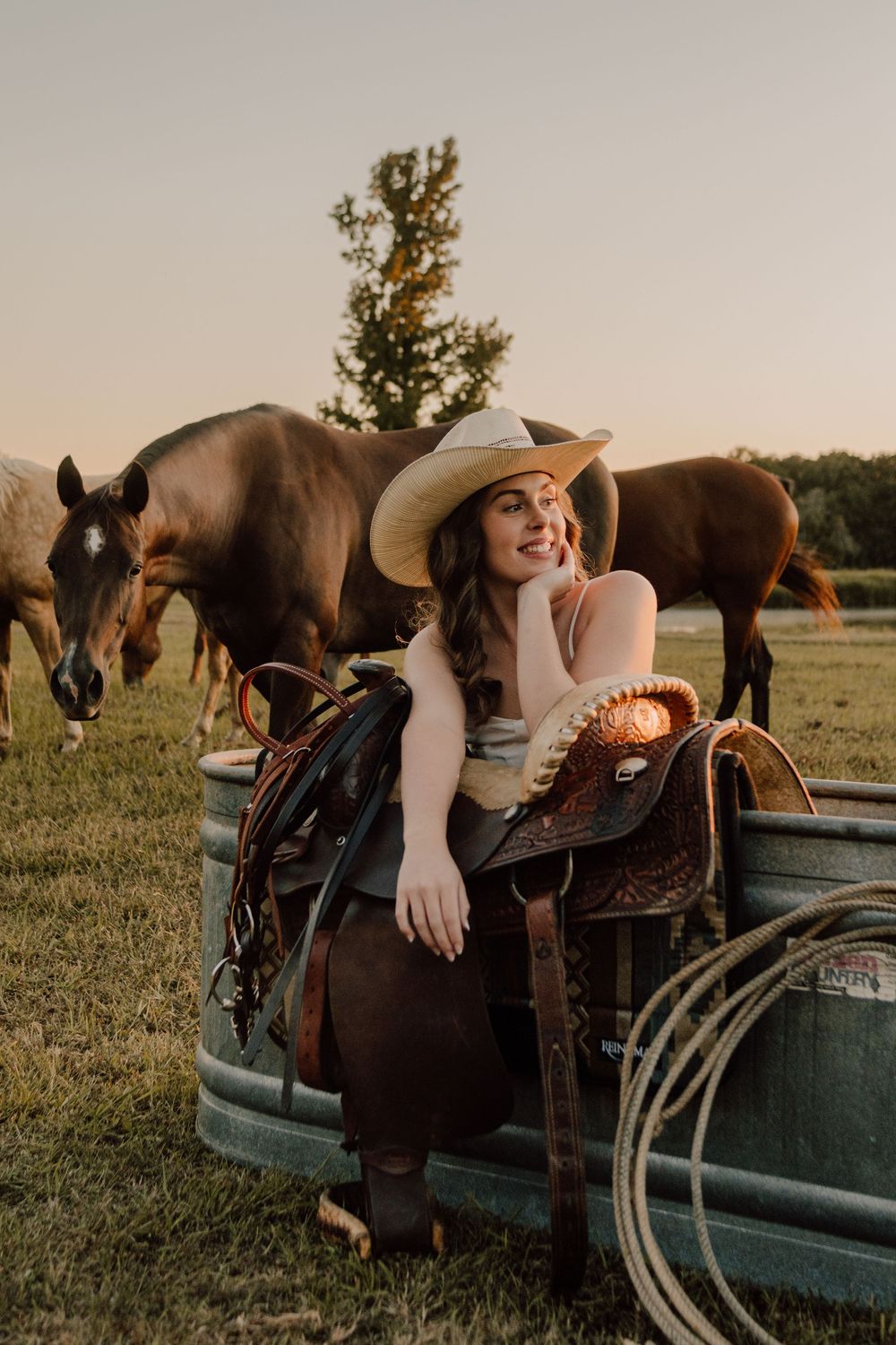 A western-style scene at sunset with horses grazing near a rustic truck while someone sits on vintage saddle equipment.