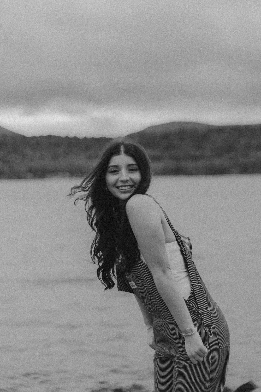 Black and white photo of someone in denim overalls smiling and leaning forward by a lake with mountains in background.