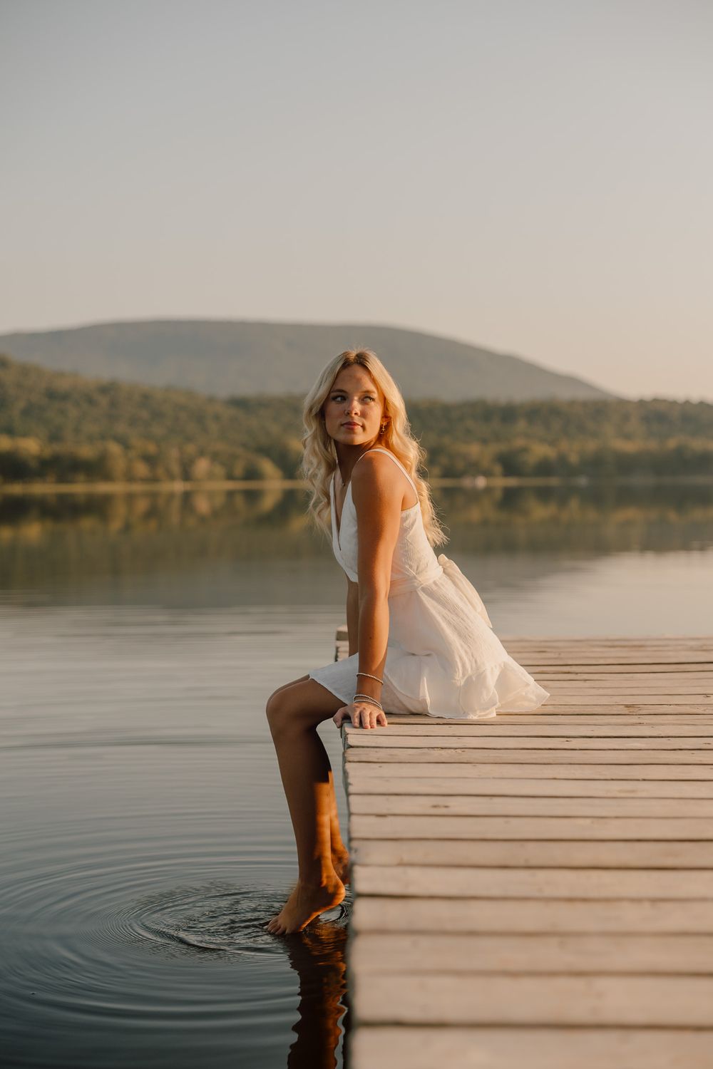 Person in white dress sits on wooden dock by serene lake at sunset with mountains in background.