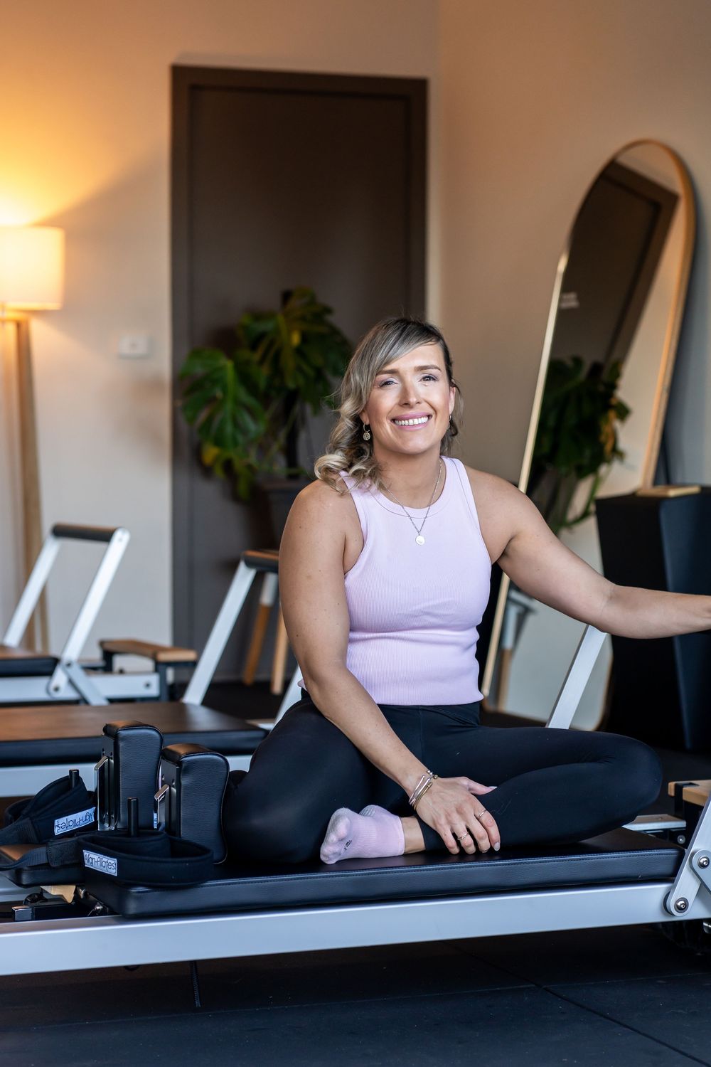 Person exercising on a Pilates reformer machine in a sunlit studio with indoor plants.