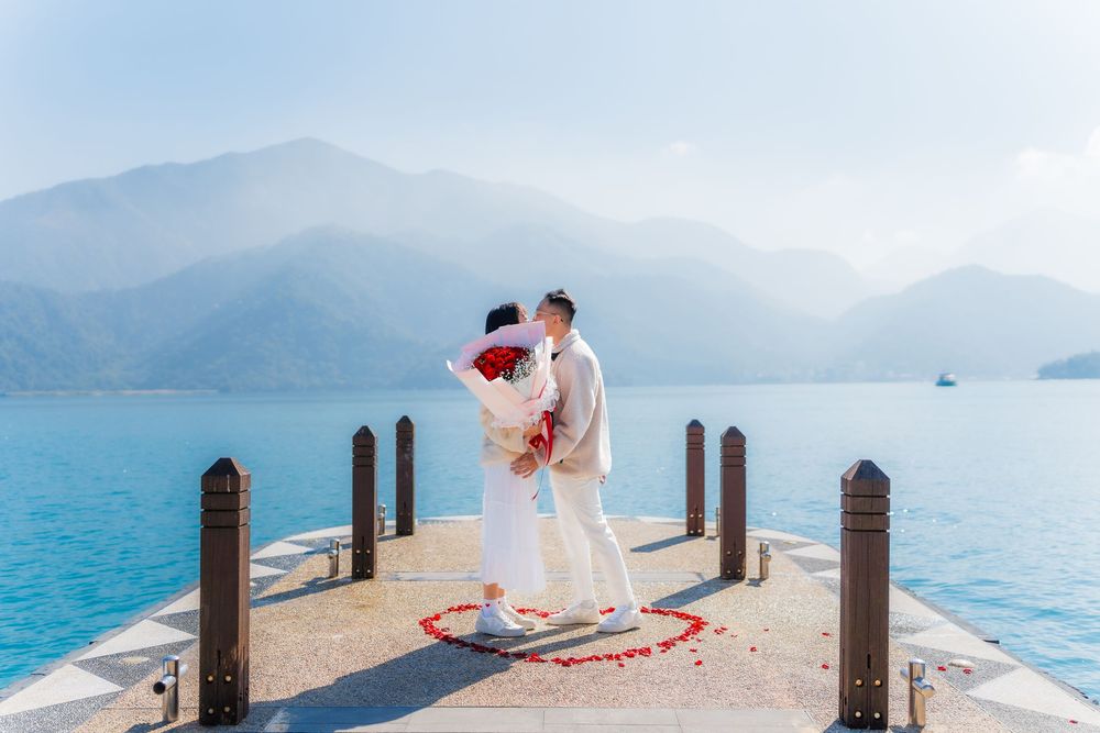A couple standing in a rose petal heart on a small pier that extends over tranquil turquoise lake with serene misty mountains landscape.