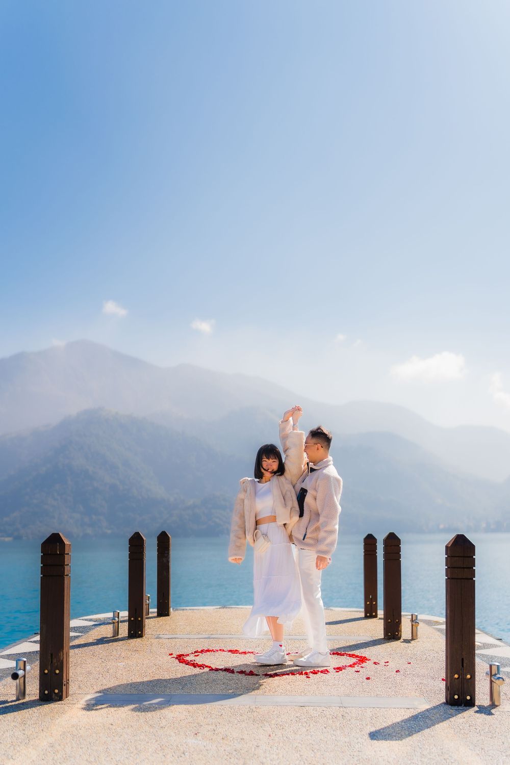 A romantic couple poses on a  pier overlooking a stunning mountain lake with blue waters and misty peaks at Sun Moon Lake, Taiwan.