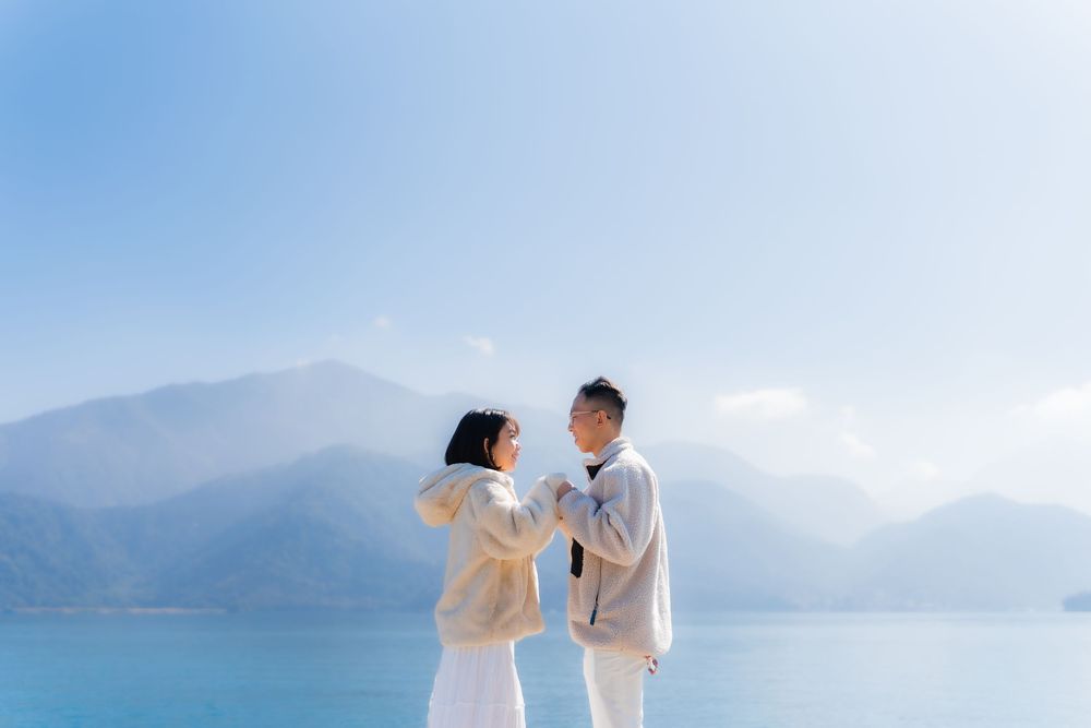 A couple stands together against a misty backdrop of indigo mountains on a bright sunny day at Taiwan's Sun Moon Lake.