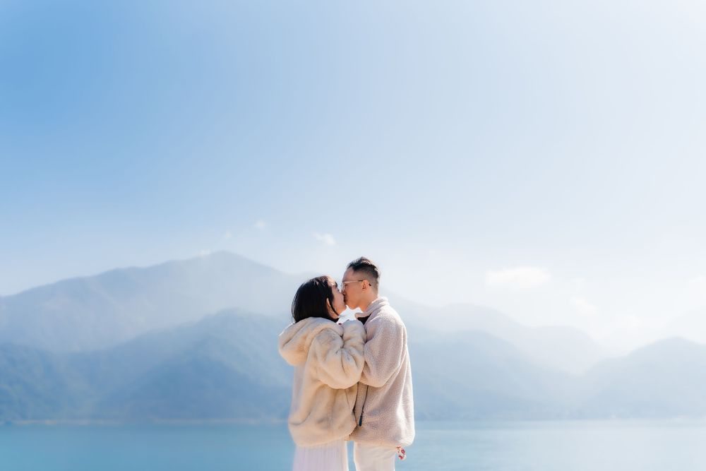 A couple in beautiful white attire share a kiss against a misty mountain backdrop near serene lake.