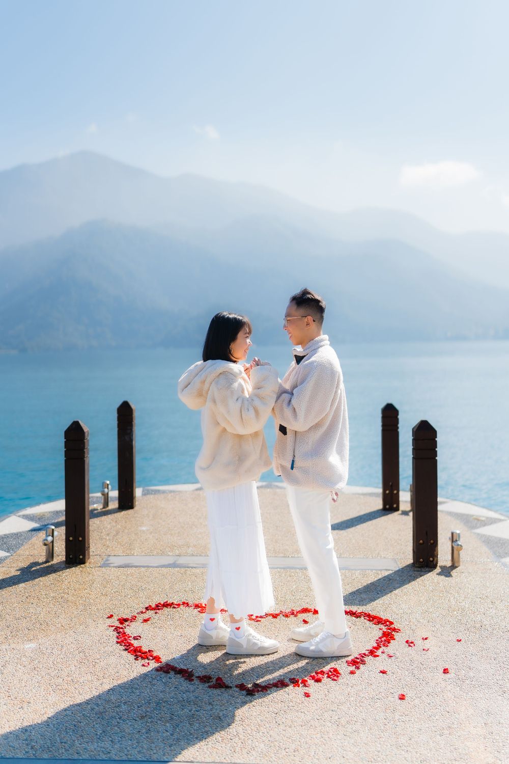 A couple in white outfits dances together on a  with mountains and water in the background, surrounded by rose petals.