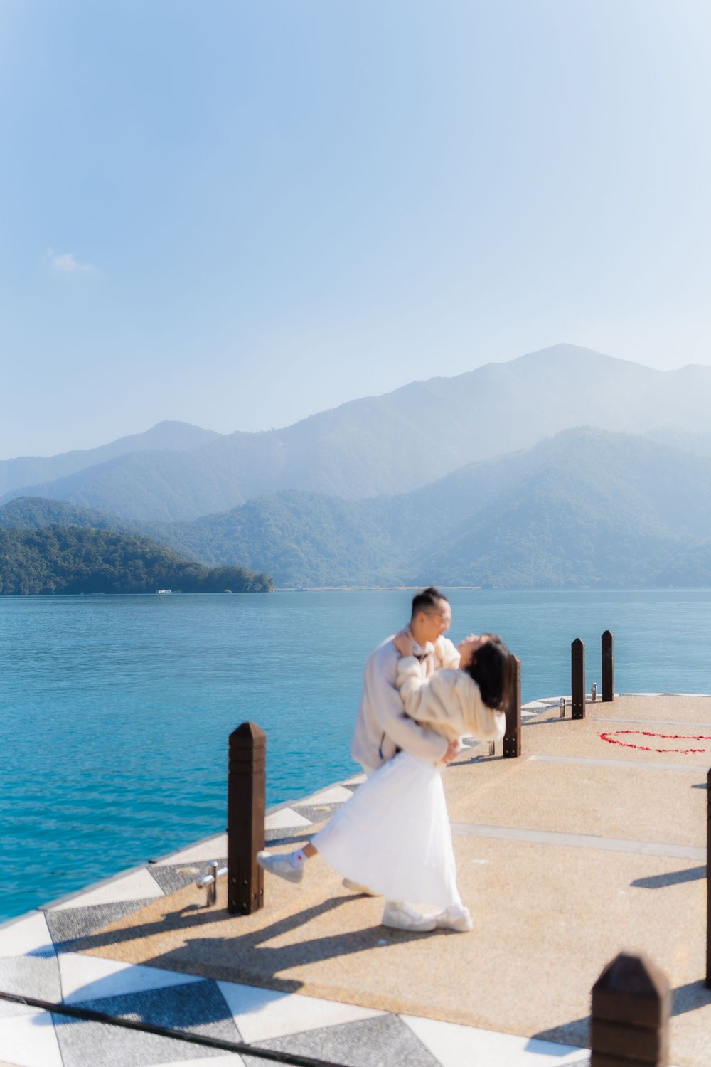 A panoramic view of a couple in white dancing on a dock extending into turquoise waters with mountains in the background on a sunny day.