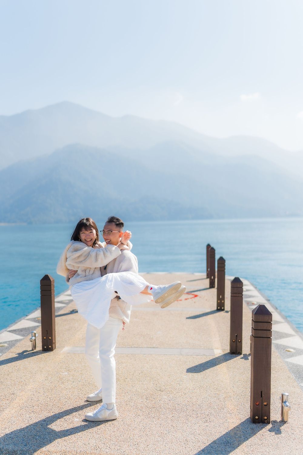 A man lovingly carries his partner sharing a romantic moment on a mountainside lake dock with misty mountain in the background.