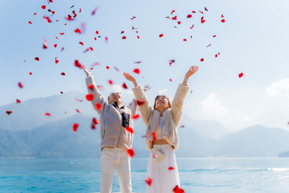 Red rose petals fall from the sky as a couple celebrates on a scenic mountain lakefront.