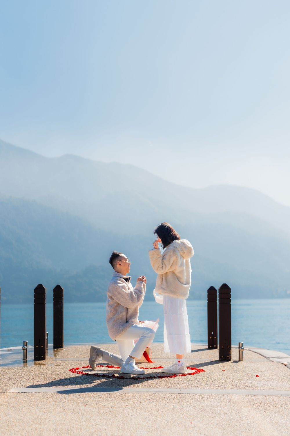 A romantic proposal scene unfolds on a lakeside dock with misty mountains and bright blue water in the background.