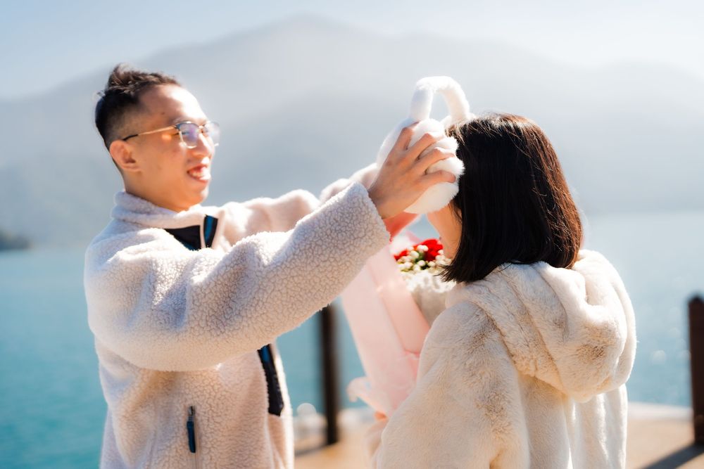 A boyfriend playfully places white fluffy earmuffs on his girlfriend near a beautiful mountain lake in the glowing morning light.