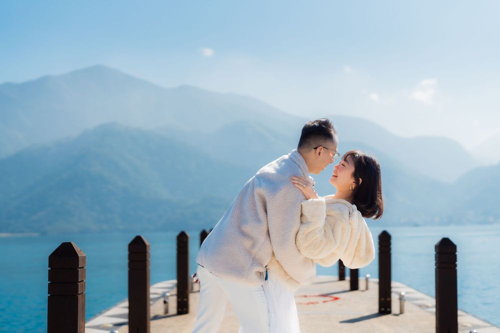 A couple in white attire embracing on a pier overlooking misty blue mountains and turquoise lake in central Taiwan.