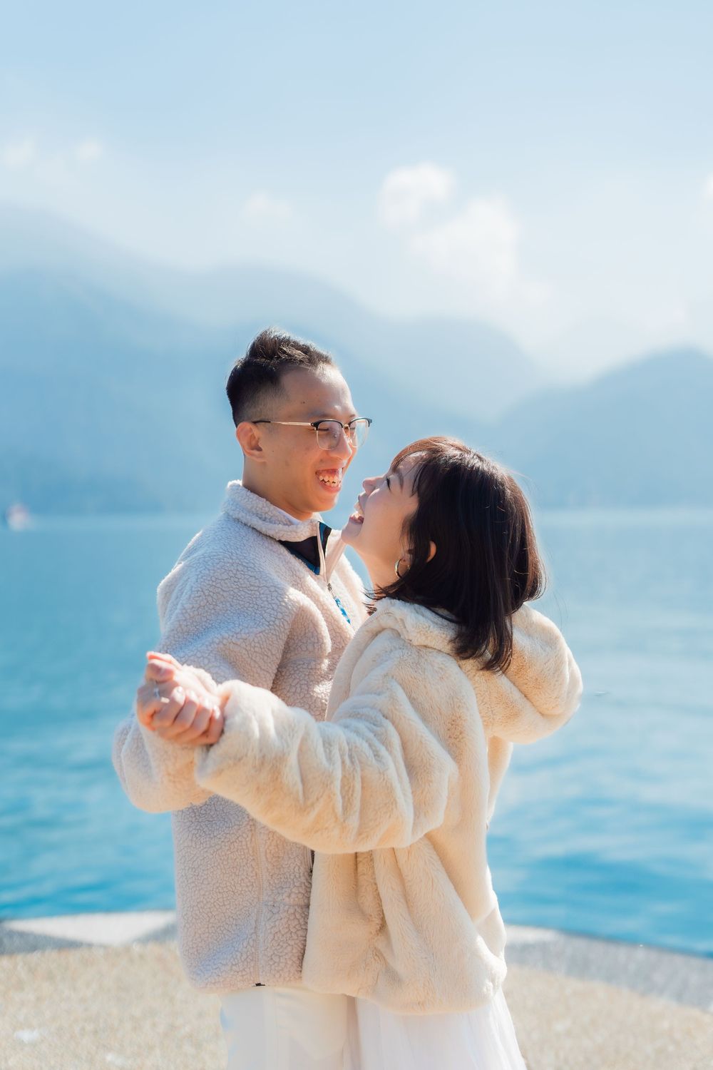 A couple wearing matching beige coats embrace and dance together by a scenic mountain lake on a bright day.