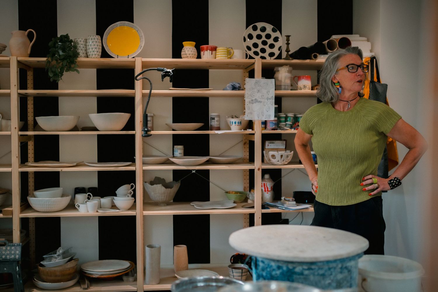 Ceramic artists working at wooden shelves filled with pottery and handmade items in a bright studio space.