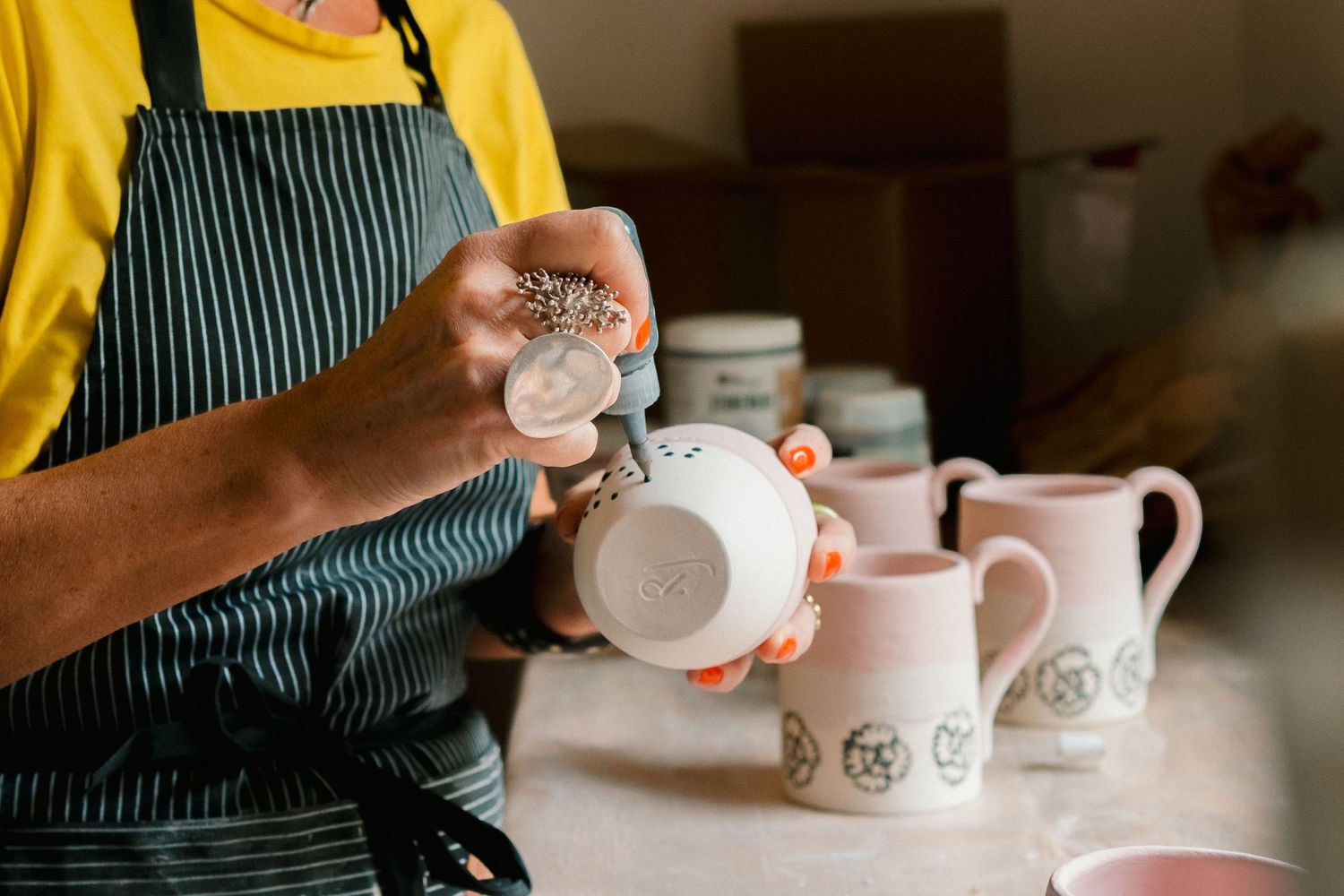 Hands in yellow sleeves pour hot water from a kettle into a white ceramic teapot on a wooden surface.