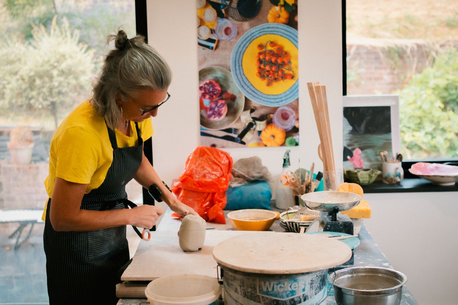 Hands working with clay on a pottery wheel in a bright studio with colorful artwork displayed on the walls in the background.