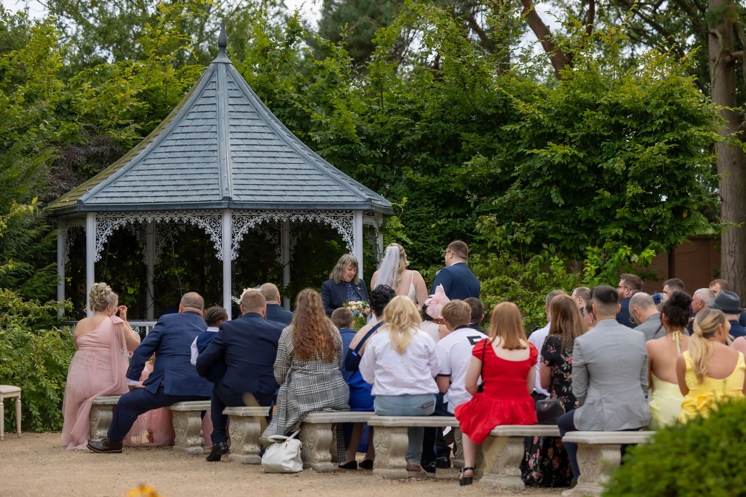 Wedding guests gather around a white gazebo in a garden setting during an outdoor ceremony.