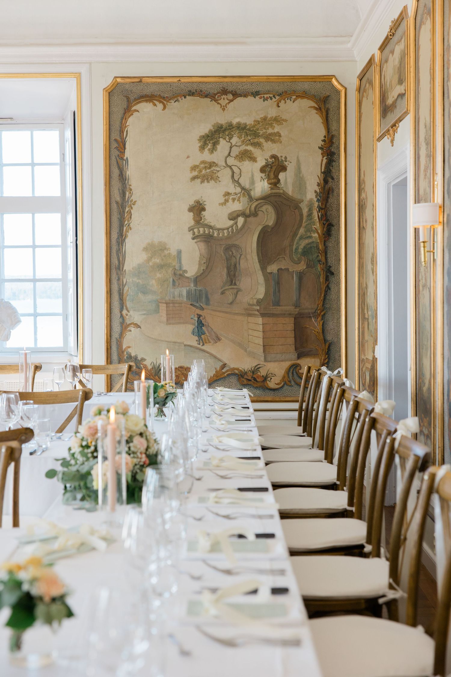 Elegant dining table with white lace tablecloth, black bistro chairs, and floral centerpiece against mural-adorned wall.