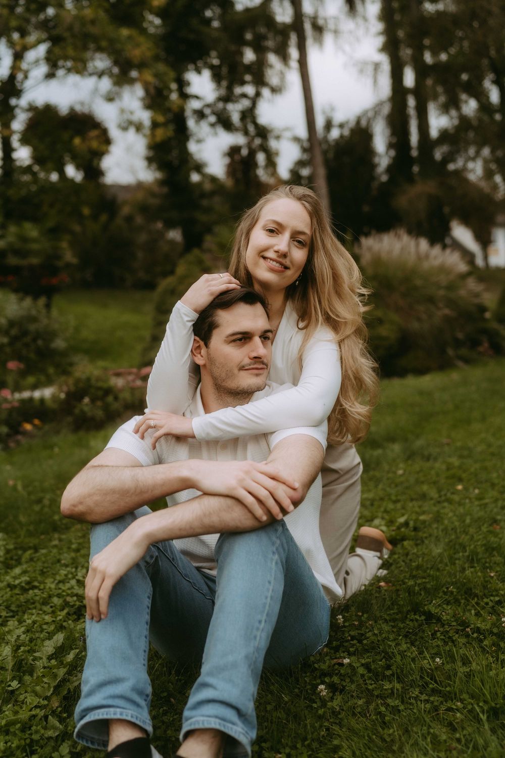 Romantic couple in white tops and jeans embrace while sitting on grass in a park setting.