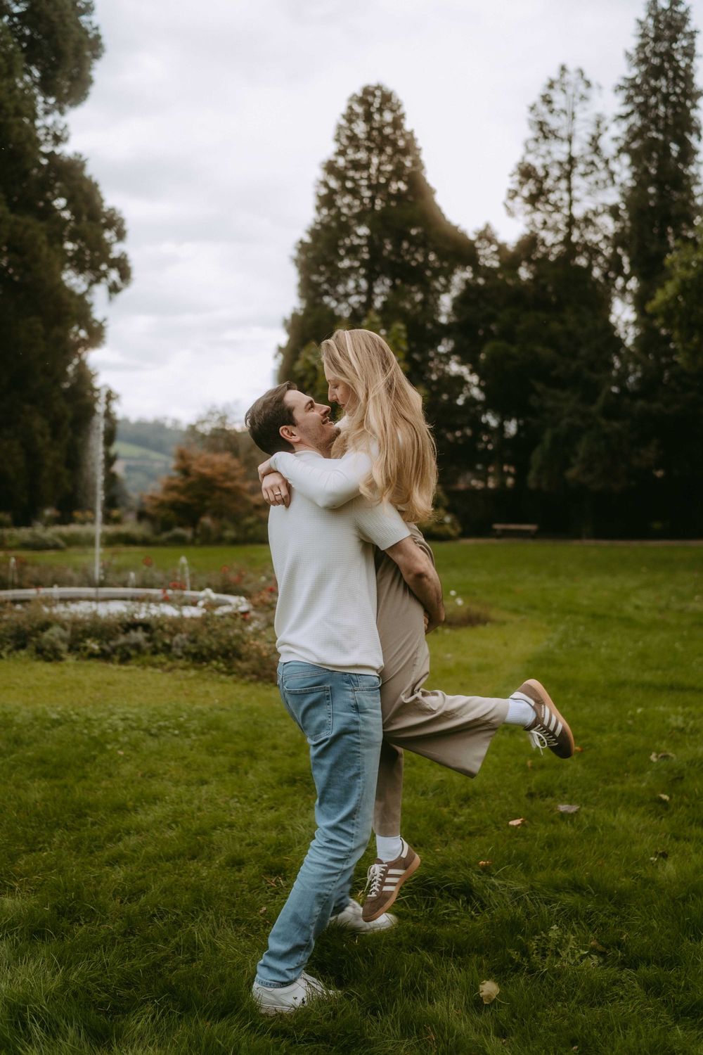 Playful couple in white sweaters and jeans dancing and embracing in a grassy field.
