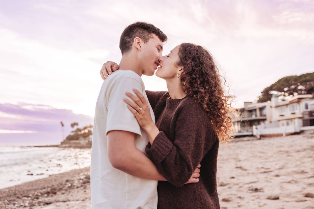 A couple shares a romantic moment on the beach at sunset with coastal homes visible in the background.
