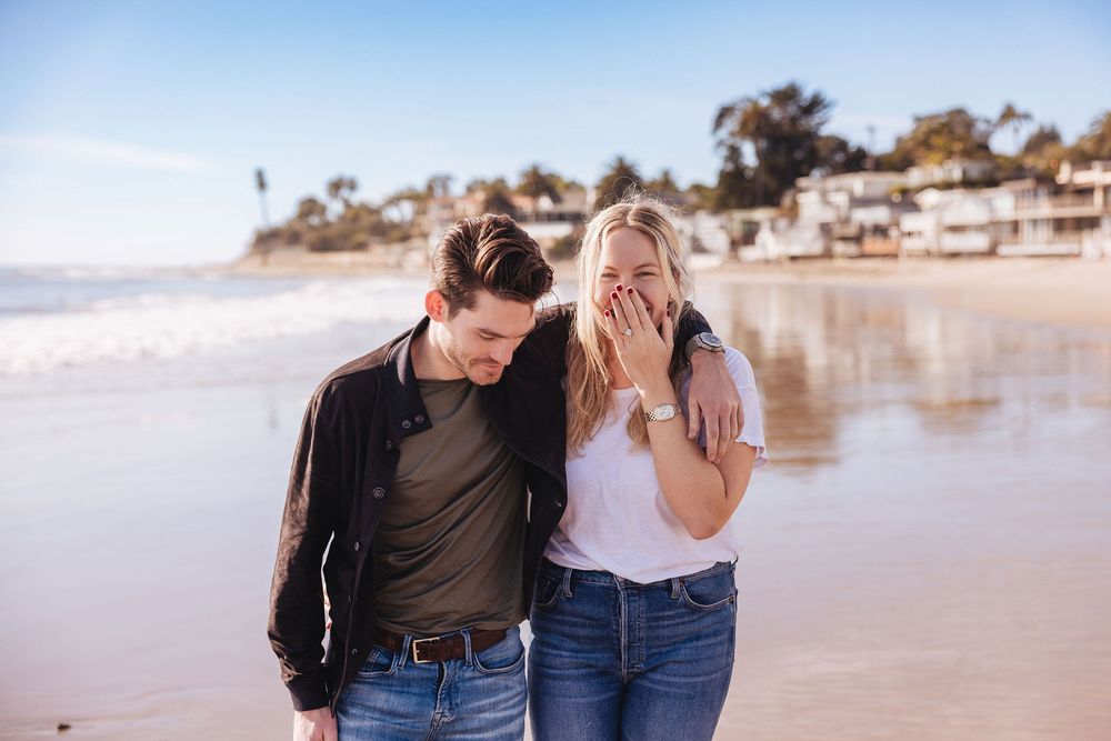 Two people share a candid laughing moment together while walking on a sunny beach with coastal homes in background.