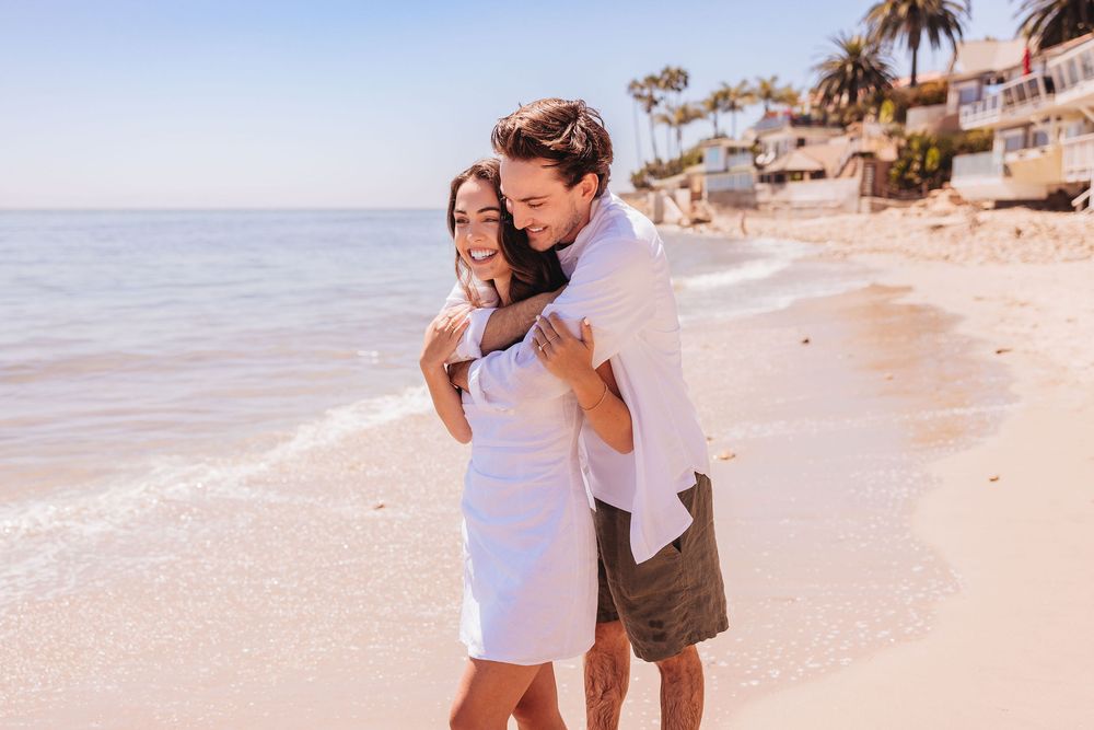 Couple in white and brown outfits walking along a sunny tropical beach with palm trees and ocean views during Santa Barbara engagement photo