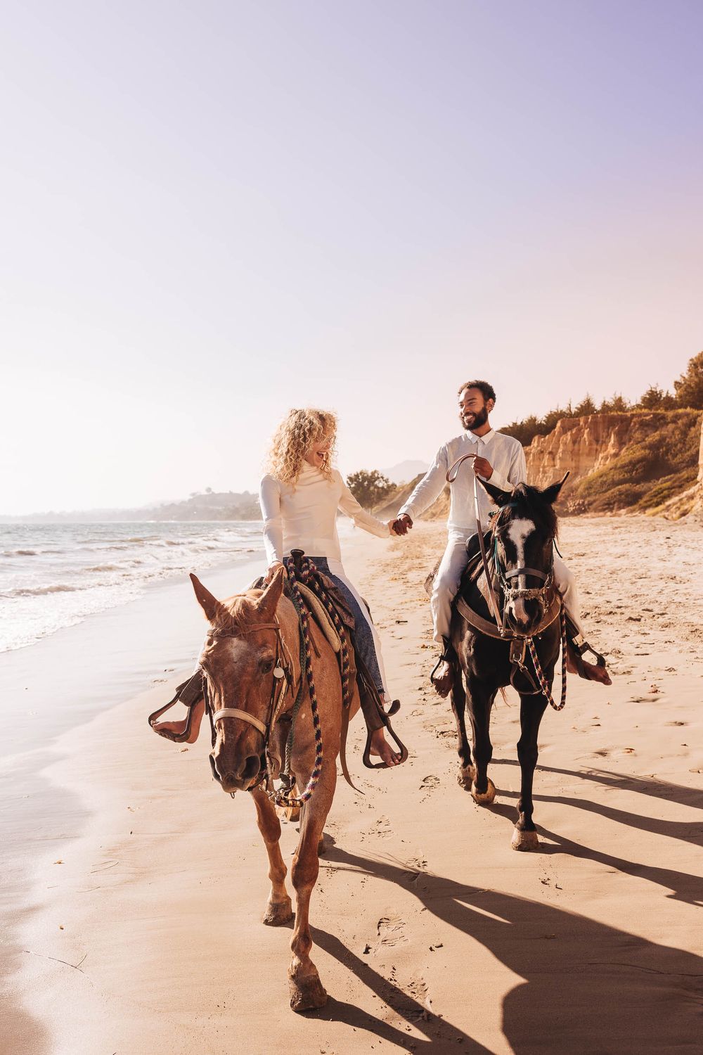 Couple riding horses along a scenic beach shoreline during sunset, casting long shadows on the sand.