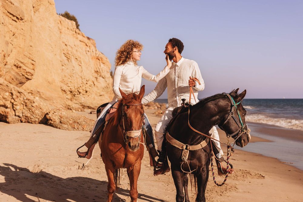 Horseback riders traverse a scenic coastal beach with towering orange cliffs and blue ocean waters in the background.