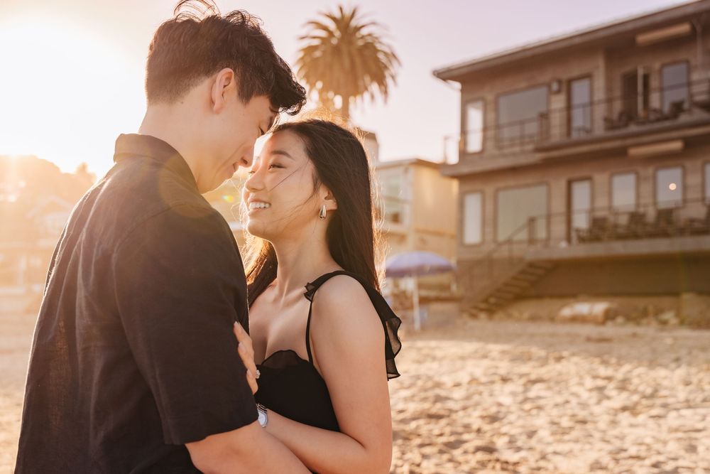 Romantic couple embraces on beach during golden hour sunset with coastal homes in background.