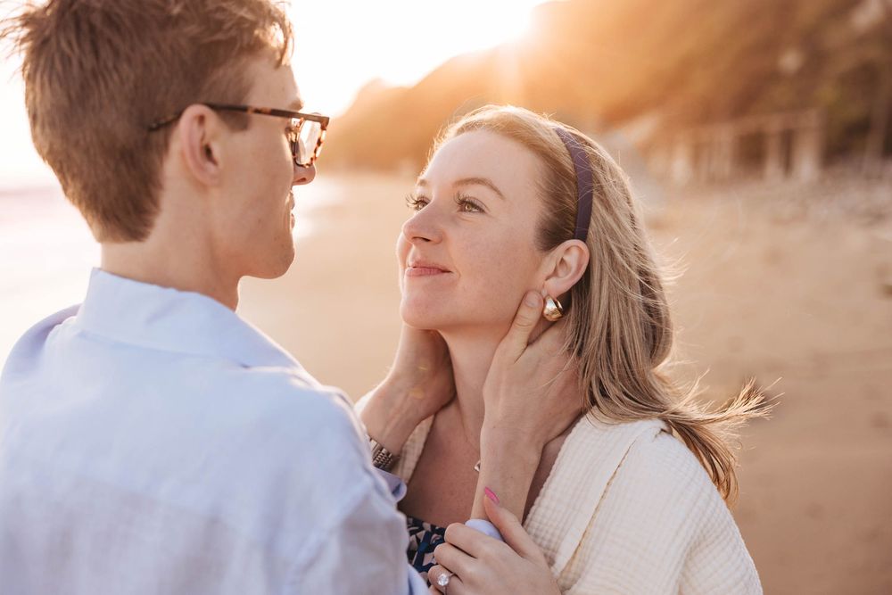 Romantic couple embraces on beach at golden hour with sun flare creating dreamy atmosphere.