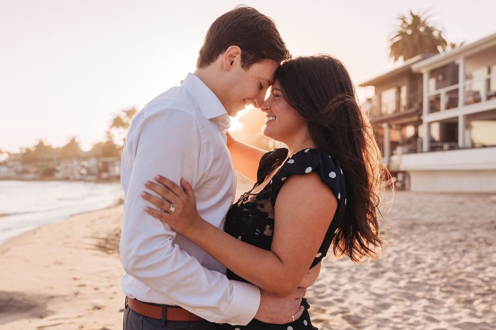 A couple embraces romantically on a sandy beach at sunset while wearing white and black casual attire.
