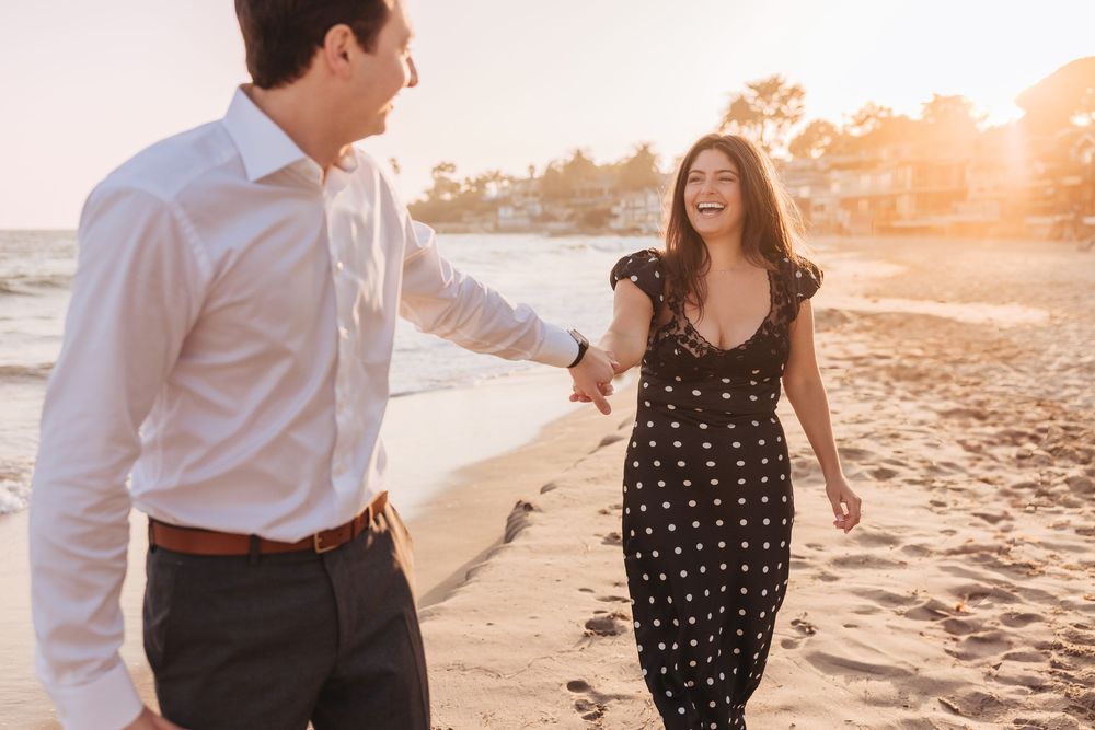 A couple walks together on a sandy beach at sunset while holding hands and smiling.